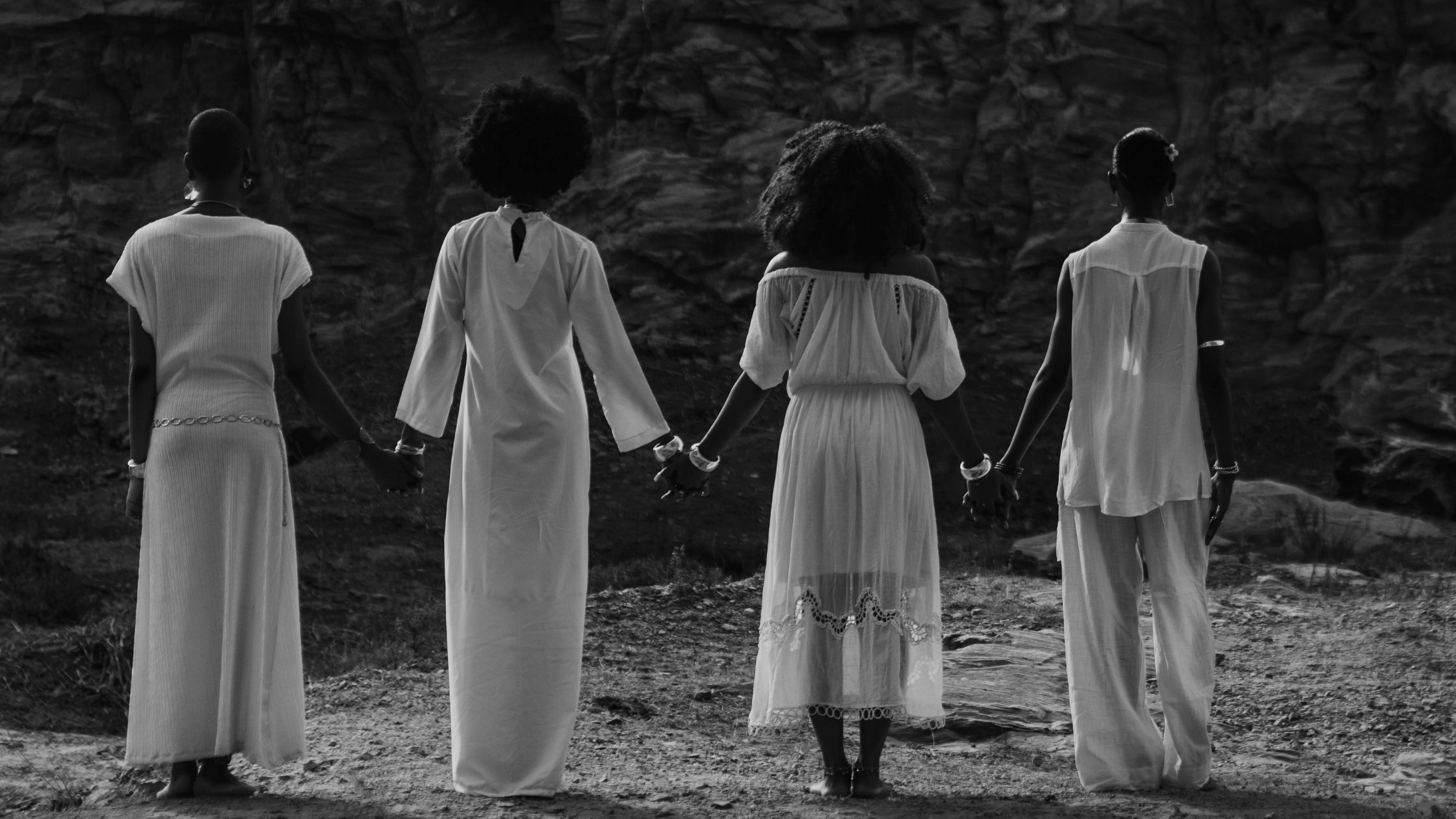 Four Black women in white clothing standing hand in hand against a dark natural backdrop.